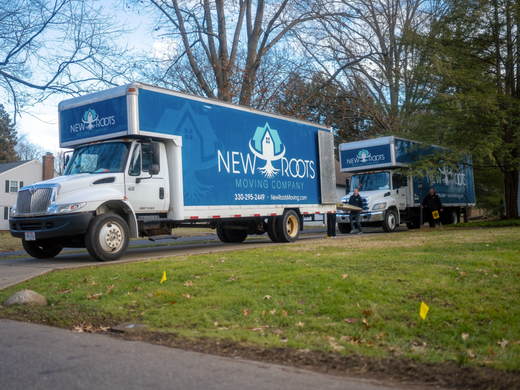 Two moving trucks from New Roots Moving Company parked on a residential street, ready for a moving job. Three team members are seen in the background, standing near the trucks, prepared for the moving process.